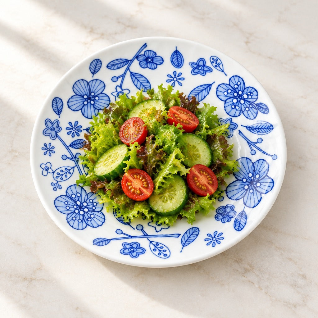 A round white ceramic plate with a blue floral pattern, filled with a green salad of lettuce, cucumber slices, and cherry tomatoes, set on a light marble surface.