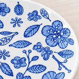 A close-up of a white ceramic plate decorated with a blue hand-painted floral and leaf pattern, set against a light wood background.