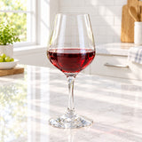 A clear wine glass with a wide bowl, partially filled with red wine, standing on a light marble kitchen countertop with a bright kitchen background.