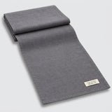 A folded dark gray woven textile, likely a blanket, throw, or scarf, displayed diagonally on a light gray background with a small white fabric label visible in the corner.