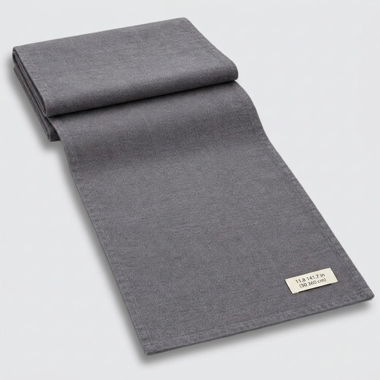 A folded dark gray woven textile, likely a blanket, throw, or scarf, displayed diagonally on a light gray background with a small white fabric label visible in the corner.