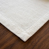 Close-up of a light beige woven textile corner with a stitched border, placed on a dark brown wooden surface.