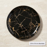 A round black plate with a gold marble pattern, shown from a top-down view on a light beige textured surface. A small label in the bottom right corner reads '3.