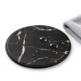 A round black faux-leather coaster with a white marble pattern, shown on a white background with water droplets on its surface and a folded gray cloth in the corner.