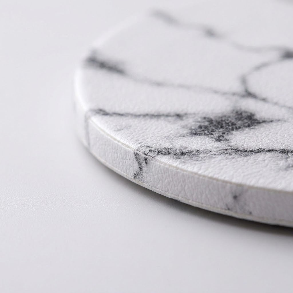 A close-up of a round white coaster with a black marble pattern, showing its textured surface and curved edge on a light gray background.