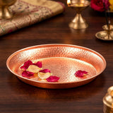 A hammered copper tray with a warm metallic finish, styled on a dark wooden table with scattered rose petals. The background features softly blurred traditional brass lamps and a patterned textile.