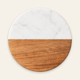 A round serving board or tray with a split design, featuring a white marble top half and a warm brown wood grain bottom half, centered on a light cream background.