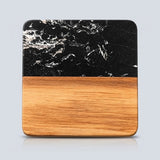A square wooden serving board or tray with rounded corners, featuring a black marble top half with white veining and a natural light brown wood grain bottom half, shown on a light gray background.
