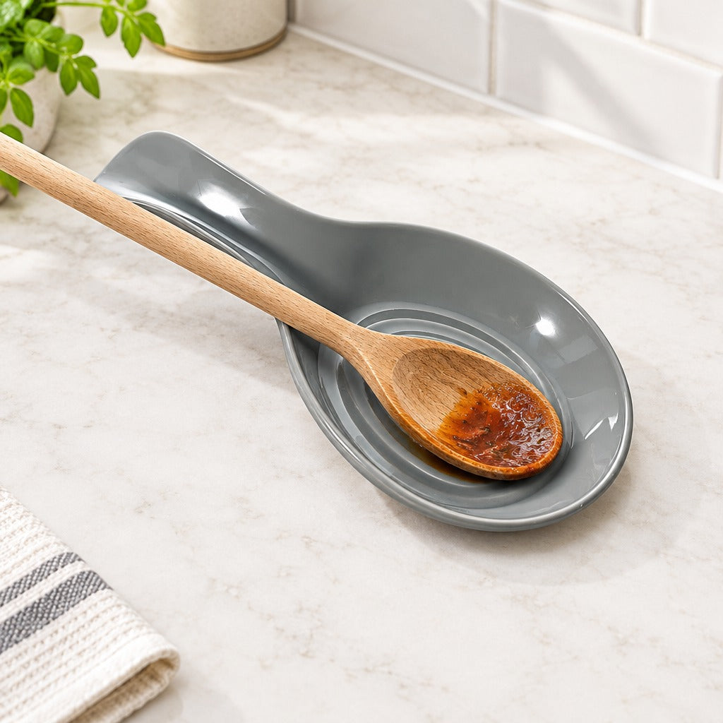 A gray ceramic spoon rest with a wooden spoon resting inside it on a light marble countertop, with a tiled backsplash and small plant in the background.