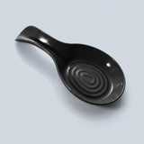 A glossy black ceramic spoon rest with a textured concentric ring design inside the bowl, shown on a light gray background.