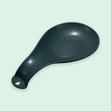 A matte black ceramic spoon rest with a hanging loop at the handle, shown on a pale mint green background.