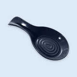 A glossy black ceramic soup spoon rest with a concentric ripple design inside the bowl, shown on a light blue background.