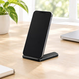 A black smartphone stand holding a black smartphone, placed on a light wood desk in a bright room with a potted plant and a closed laptop nearby.