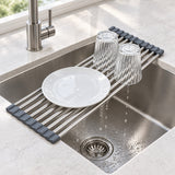 A stainless steel sink drain guard with dark gray silicone end caps, shown in a kitchen sink with a white plate and two clear drinking glasses resting on it, with water dripping into the drain.