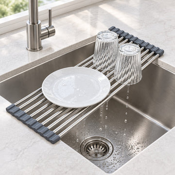 A stainless steel sink drain guard with dark gray silicone end caps, shown in a kitchen sink with a white plate and two clear drinking glasses resting on it, with water dripping into the drain.