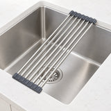 A stainless steel sink drain guard with dark gray rubber end caps, shown resting across a stainless steel kitchen sink.