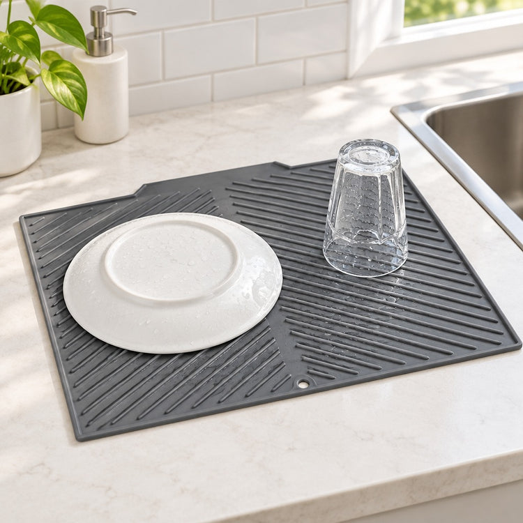 A dark gray silicone dish drying mat on a light marble kitchen counter, shown with a white plate and a clear glass cup placed on it near a sink.