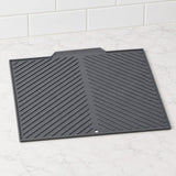 A black silicone kitchen mat with a diagonal ribbed pattern, shown on a white marble countertop with white subway tiles in the background.