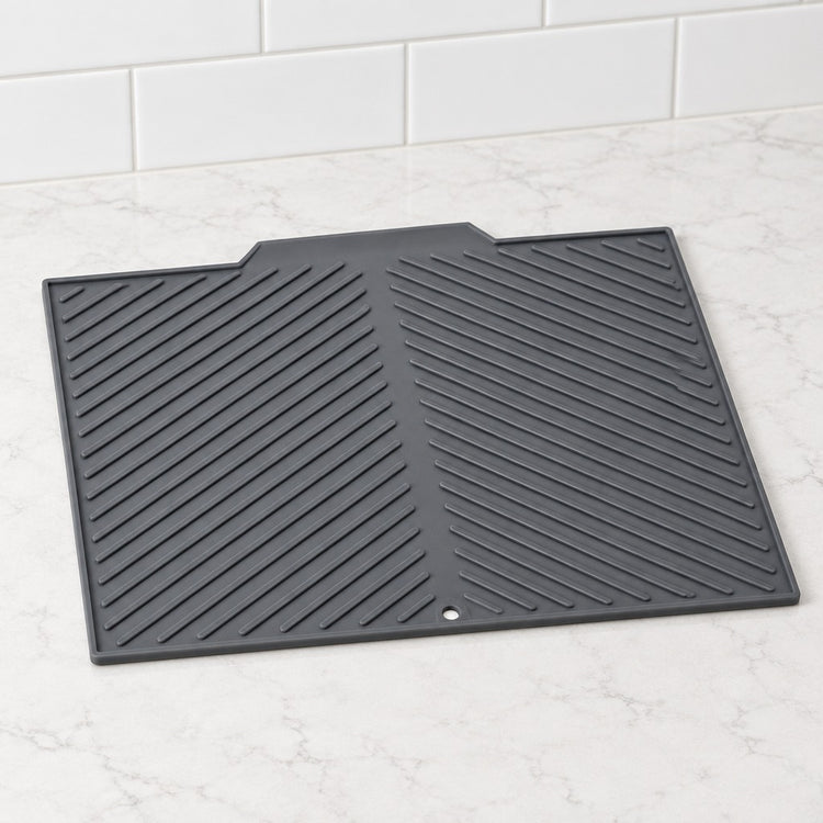 A black silicone kitchen mat with a diagonal ribbed pattern, shown on a white marble countertop with white subway tiles in the background.