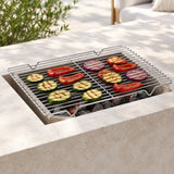 A rectangular metal grill grate sits over a built-in stone fire pit with charcoal and grilled vegetables, shown in an outdoor patio setting.