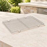 A rectangular stainless steel wire cooling rack with integrated handles, shown on a beige stone countertop outdoors with a blurred kitchen or patio background.