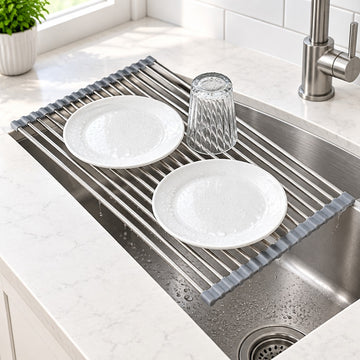 A stainless steel kitchen sink drain guard with gray rubber end caps, shown holding two white plates and a clear drinking glass in a sink. The scene is set in a bright, modern kitchen with a white marble countertop and a chrome faucet.