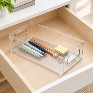 A clear plastic drawer organizer tray with metal handles, shown inside an open wooden drawer holding pens, pencils, and a sticky note pad.