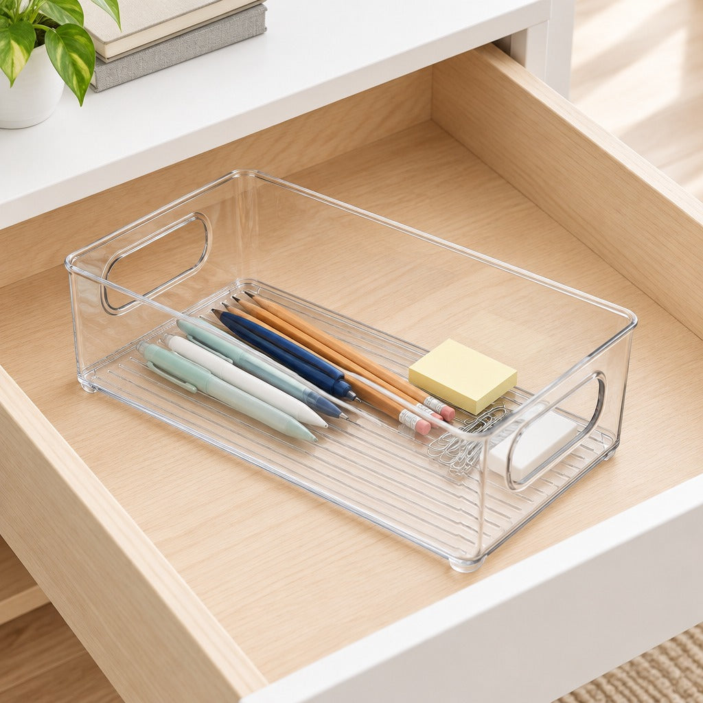 A clear plastic drawer organizer tray with metal handles, shown inside an open wooden drawer holding pens, pencils, and a sticky note pad.