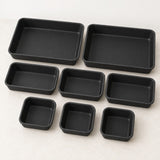 A set of eight black rectangular non-stick baking pans in various sizes, arranged neatly on a light beige surface.