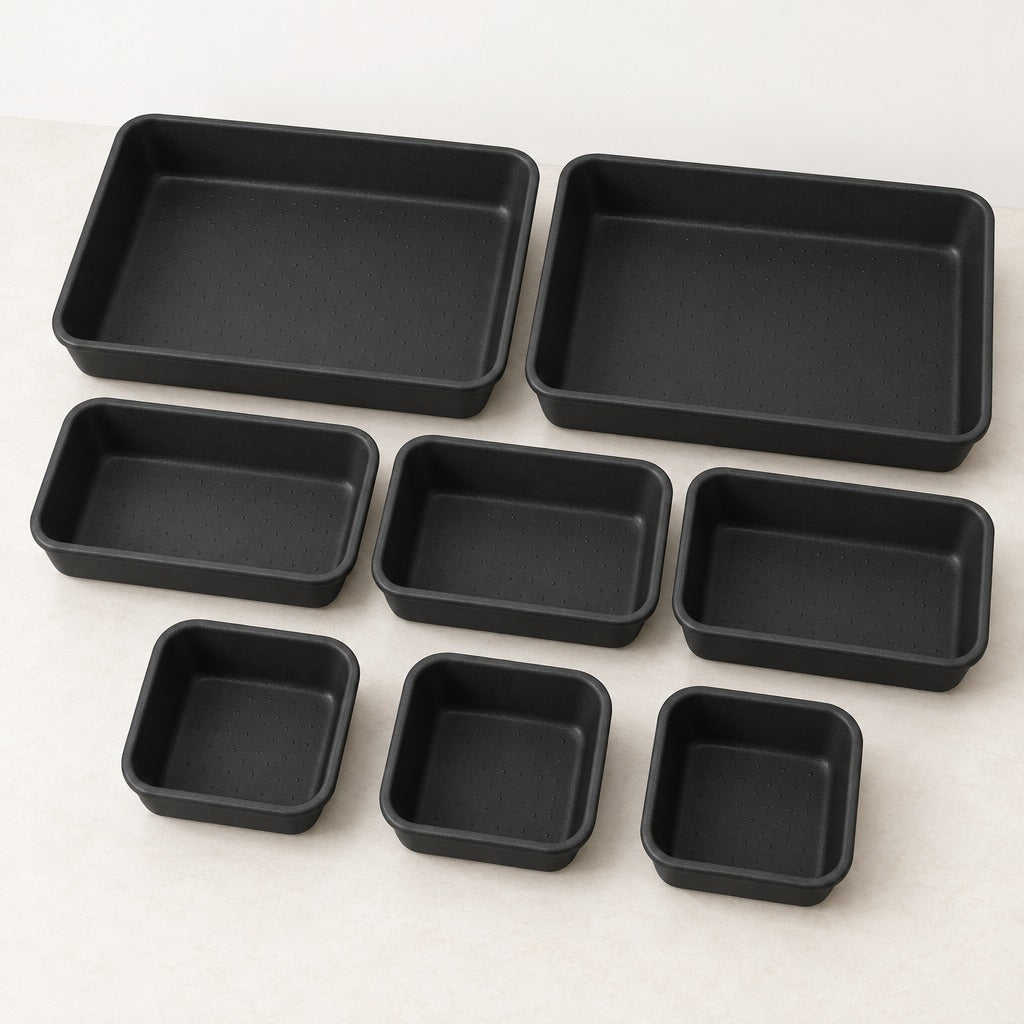 A set of eight black rectangular non-stick baking pans in various sizes, arranged neatly on a light beige surface.