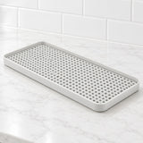 A rectangular white silicone kitchen draining tray with a raised rim and a grid of small drainage holes, shown on a white marble countertop with a white subway tile backsplash.