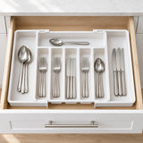A white plastic cutlery organizer tray arranged inside an open kitchen drawer, holding silver metal utensils including spoons, forks, knives, and a spoon resting on top.