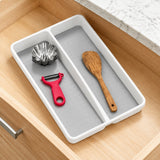 A white plastic drawer organizer tray with two compartments, shown inside an open wooden drawer. The tray contains kitchen utensils, including a metal flower-shaped cutter, a red peeler, and a wooden spoon, on a marble countertop.