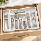 A white plastic drawer organizer tray holding a set of stainless steel flatware, including forks, spoons, and knives, arranged inside an open wooden drawer.