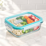 A clear glass food storage container with a bright teal lid, shown filled with rice, broccoli, carrots, and sliced strawberries on a light marble surface with a white tile backsplash in the background.