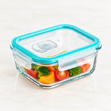 A square clear glass storage container with a bright turquoise blue lid and handle, shown on a light marble-like surface with colorful vegetables inside.