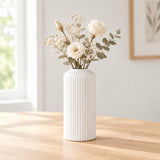 A white ribbed ceramic vase filled with dried white flowers and green eucalyptus stems, placed on a light wood table in a softly lit room.