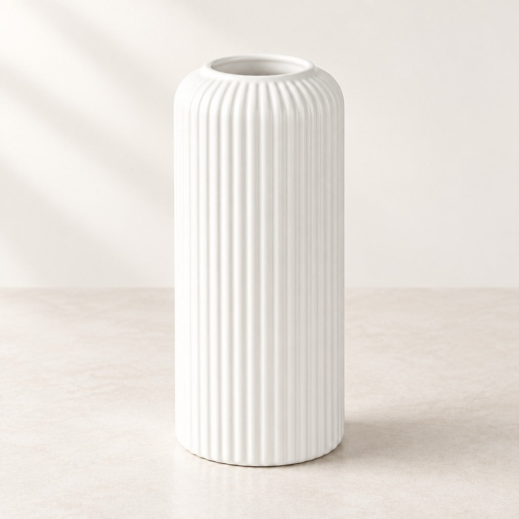 A tall white ceramic vase with a fluted vertical ribbed texture, standing on a light beige surface against a neutral cream background.