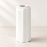 A tall white ceramic vase with a fluted vertical ribbed texture, standing on a light beige surface against a neutral cream background.
