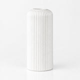 A white ceramic vase with a fluted, vertically ribbed cylindrical design, standing on a reflective white surface against a seamless white background.