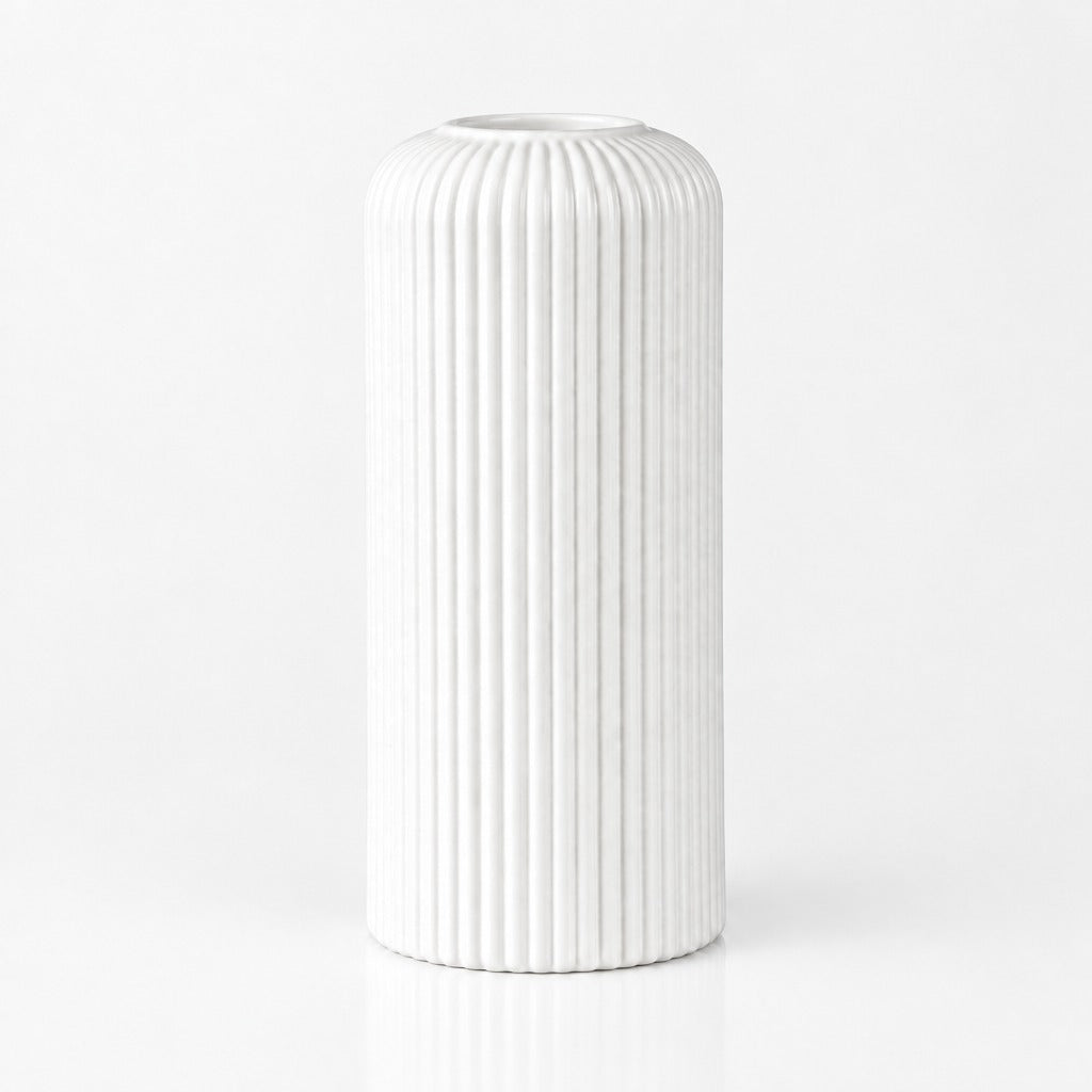 A white ceramic vase with a fluted, vertically ribbed cylindrical design, standing on a reflective white surface against a seamless white background.