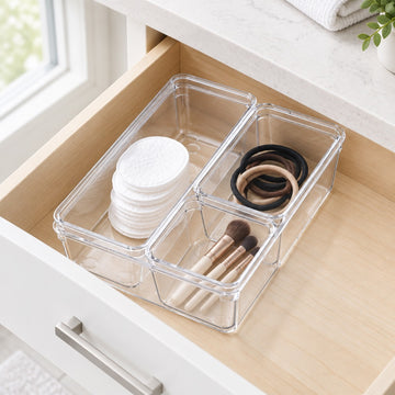 Clear plastic drawer organizer bins arranged inside an open wooden drawer, holding cotton pads, makeup brushes, and hair ties.