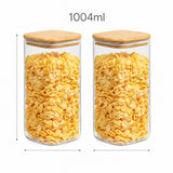 Two clear glass canisters filled with yellow corn flakes, shown side by side on a white background with measurement arrows and the text '1004ml' above. The canisters have light wood lids.