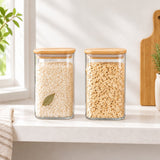 Two clear glass canisters with light wood lids sit on a white countertop in a bright kitchen setting. The left canister is filled with beige grains or pasta, and the right canister is filled with flatbread or crackers.