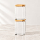 Two clear glass storage jars with light natural wood lids, stacked vertically on a light beige marble surface against a neutral off-white background.