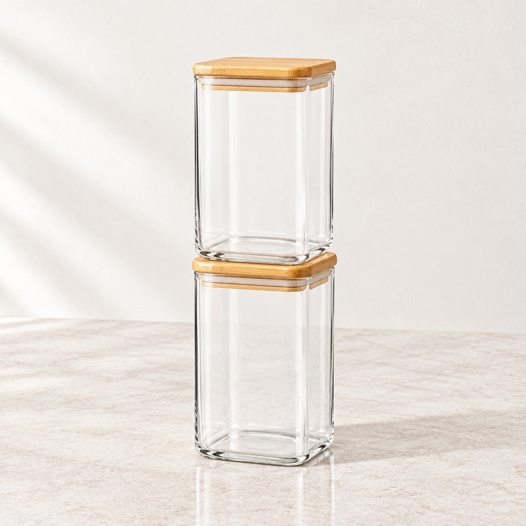 Two clear glass storage jars with light natural wood lids, stacked vertically on a light beige marble surface against a neutral off-white background.