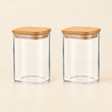 Two clear glass canisters with square bamboo lids, shown side by side on a light beige surface against a matching neutral background.