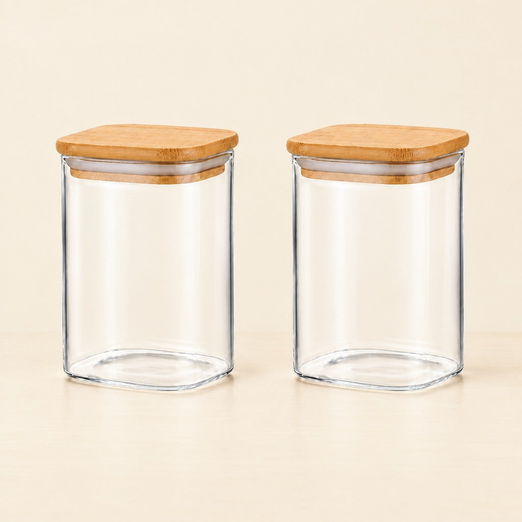 Two clear glass canisters with square bamboo lids, shown side by side on a light beige surface against a matching neutral background.