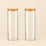 Two tall, clear glass canisters with light brown bamboo lids, standing side-by-side on a light beige surface against a matching beige background.