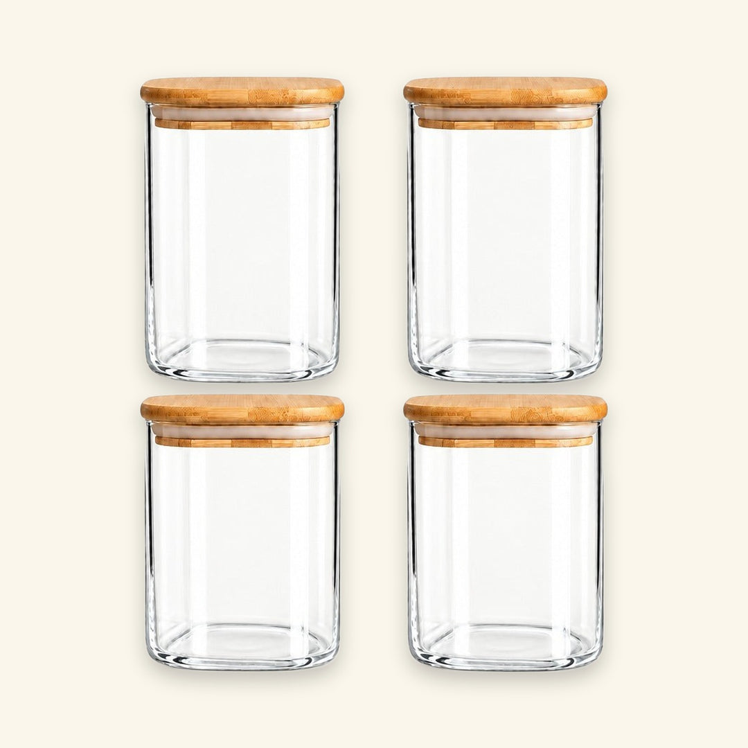 Four clear glass canisters with light brown bamboo lids, arranged in a 2x2 grid on a warm off-white background.